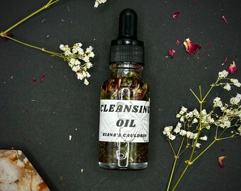 May include: A small glass bottle with a black dropper top filled with a clear liquid and dried rose petals. The bottle has a white label that reads "CLEANSING OIL KIANA'S CAULDRON".