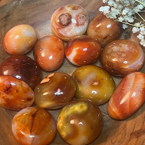 May include: A collection of carnelian tumbled stones in various shades of orange, red, and brown. The stones are smooth and polished, with some showing natural banding and inclusions.