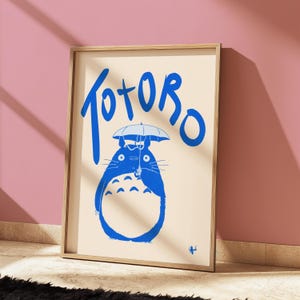 May include: A framed print featuring a blue illustration of Totoro holding an umbrella, with the word "Totoro" in blue script above. The artwork is set against a cream background and framed in light wood.