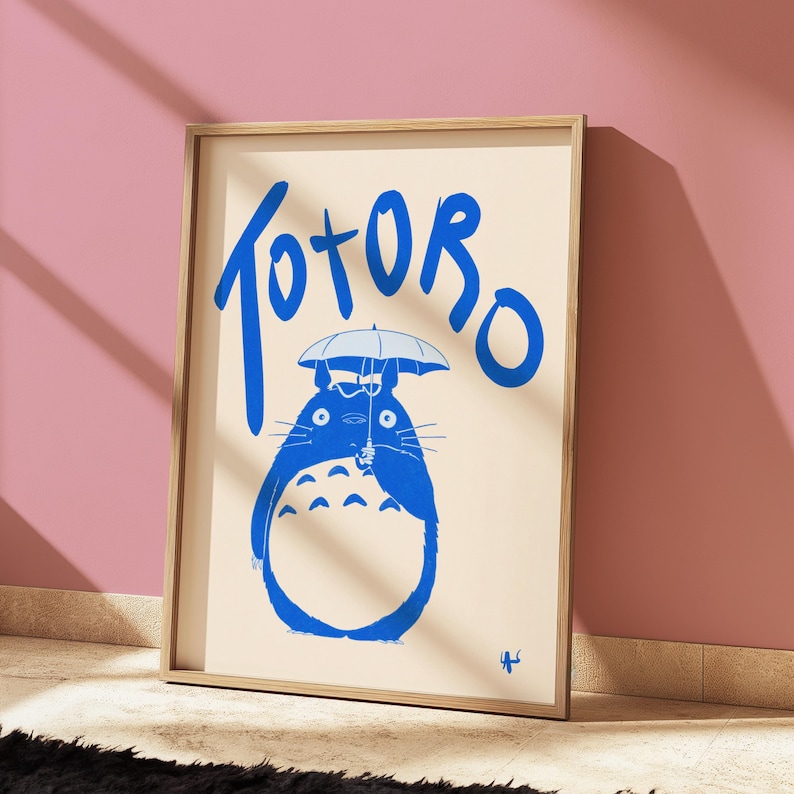 May include: A framed print featuring a blue illustration of Totoro holding an umbrella, with the word "Totoro" in blue script above. The artwork is set against a cream background and framed in light wood.