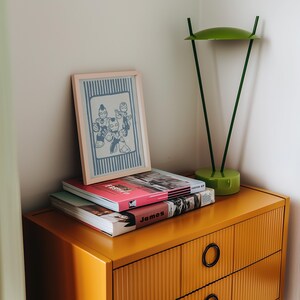 May include: A mustard yellow chest of drawers with black circular handles. A framed print with a blue and white cartoon illustration sits on a stack of books. A green desk lamp is also on the chest.
