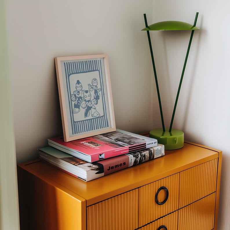May include: A mustard yellow chest of drawers with black circular handles. A framed print with a blue and white cartoon illustration sits on a stack of books. A green desk lamp is also on the chest.