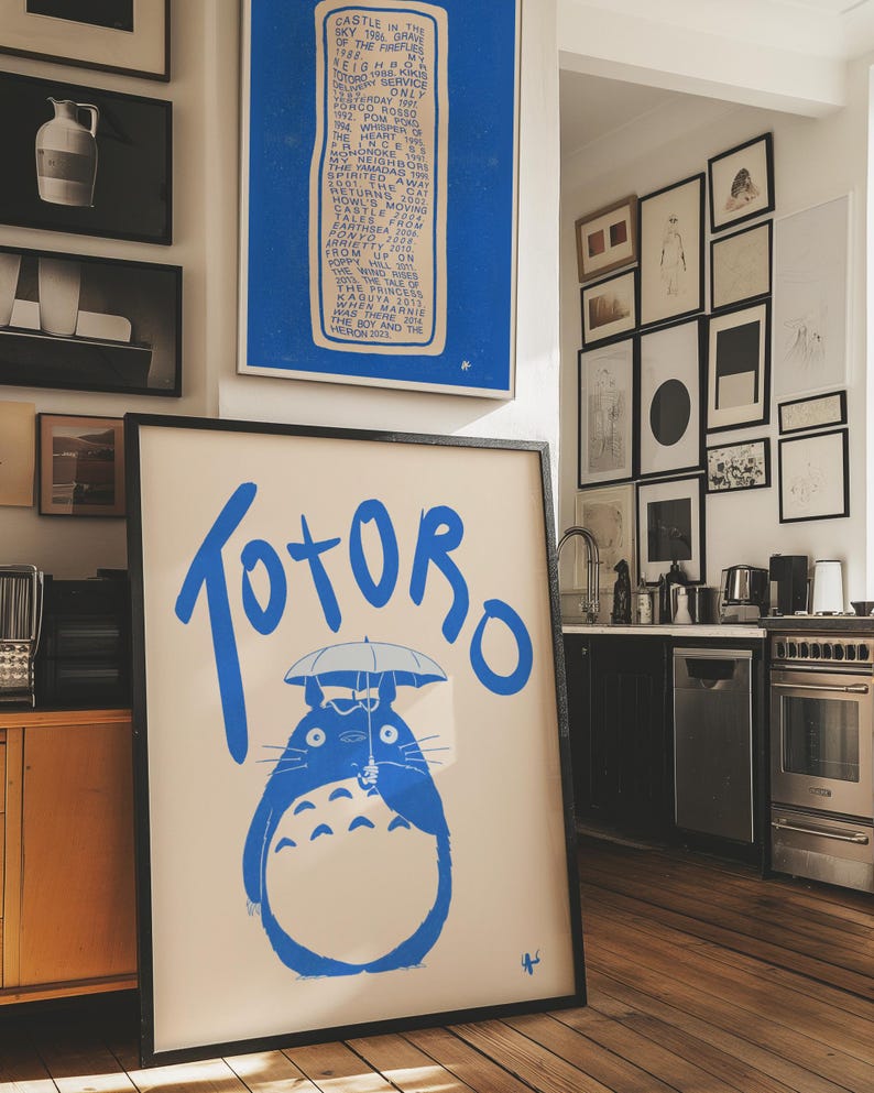 May include: A black framed poster with a cream background and blue text that says 'TOTORO' in large letters. A blue illustration of Totoro holding an umbrella is in the center of the poster. The poster is hanging on a wall in a room with a wooden floor.