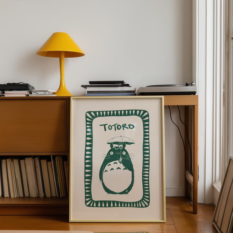 Puede incluir: Un estampado enmarcado con el personaje Totoro en verde sobre un fondo crema. El estampado est&aacute; en un marco dorado. En la habitaci&oacute;n tambi&eacute;n se ven una l&aacute;mpara amarilla, un mueble de madera y un tocadiscos.