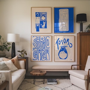 May include: Four framed art prints with blue and white designs hang on a white wall. The prints feature illustrations and text, including the word "Totoro." The room includes a sofa, chair, and wooden furniture.