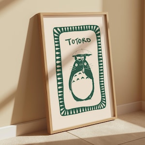 Puede incluir: Un estampado enmarcado con una ilustraci&oacute;n verde de Totoro sosteniendo un paraguas sobre un fondo crema. La palabra "Totoro" est&aacute; escrita encima del personaje. El estampado est&aacute; en un marco de madera marr&oacute;n claro.