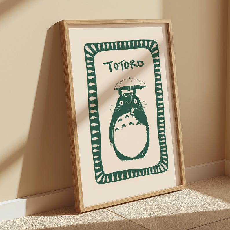 Puede incluir: Un estampado enmarcado con una ilustraci&oacute;n verde de Totoro sosteniendo un paraguas sobre un fondo crema. La palabra "Totoro" est&aacute; escrita encima del personaje. El estampado est&aacute; en un marco de madera marr&oacute;n claro.