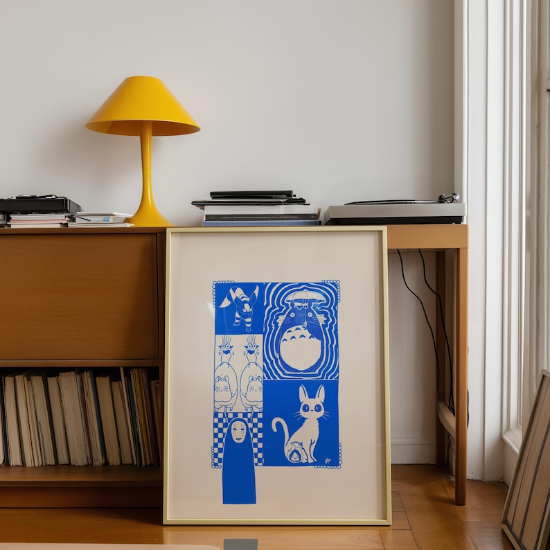 May include: A framed art print featuring blue and white illustrations of characters from the animated film, My Neighbor Totoro. The print is in a gold frame and is displayed on a wooden cabinet. A yellow lamp sits on top of the cabinet.