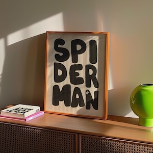May include: A framed art print with the word "SPIDERMAN" in bold, black letters. The print has an orange frame and is displayed on a wooden cabinet. A green vase and two books are also on the cabinet.