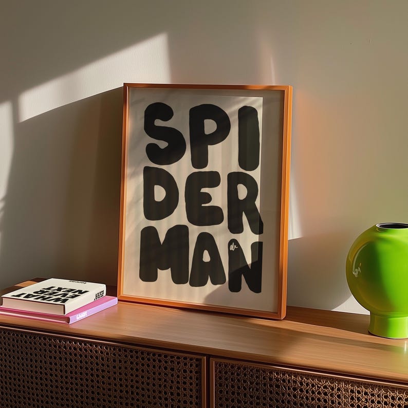 May include: A framed art print with the word "SPIDERMAN" in bold, black letters. The print has an orange frame and is displayed on a wooden cabinet. A green vase and two books are also on the cabinet.