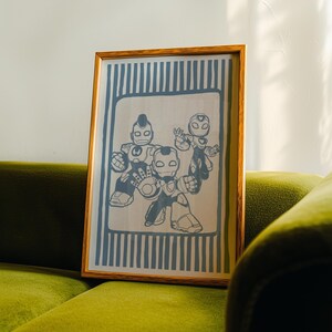 May include: A framed print depicting three cartoon figures in Iron Man-style suits. The artwork is in shades of grey and black, set against a light background with blue vertical stripes. The frame is made of wood.