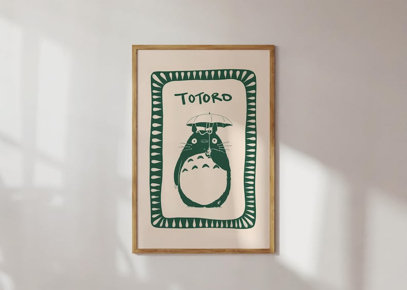 Puede incluir: Una impresi&oacute;n enmarcada con una ilustraci&oacute;n verde de Totoro sosteniendo un paraguas. La obra de arte est&aacute; sobre un fondo crema, con la palabra "TOTORO" encima del personaje. El marco es de color marr&oacute;n claro.