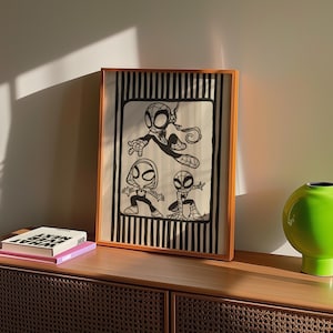 May include: Framed artwork featuring three stylized Spider-Man characters in black and white. The artwork is set against a striped background and framed in a light brown wooden frame. The artwork is displayed on a wooden cabinet.