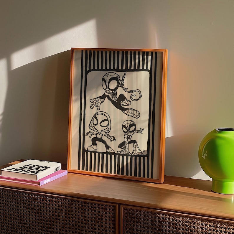 May include: Framed artwork featuring three stylized Spider-Man characters in black and white. The artwork is set against a striped background and framed in a light brown wooden frame. The artwork is displayed on a wooden cabinet.