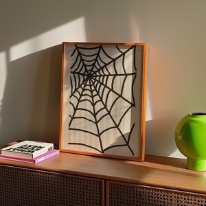 May include: A framed artwork featuring a black spiderweb design on a cream background. The artwork is encased in a warm-toned wooden frame. A green vase and books are on a wooden cabinet.