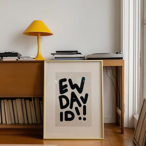Op de afbeelding: Een ingelijste kunstdruk met de tekst "EW DAVID!!" in vette zwarte letters. De print zit in een gouden lijst. Een gele lamp en andere items zijn zichtbaar op een houten tafel op de achtergrond.