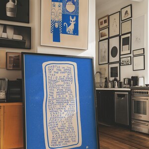 May include: A framed blue print with white text listing animated film titles and dates. Above, a framed blue and white print with cartoon characters. The image is in a room with other framed art and a kitchen.