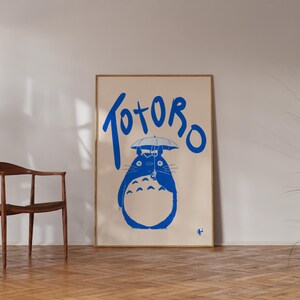 K&ouml;nnte beinhalten: Ein gerahmtes Poster mit einer blauen Illustration von Totoro mit einem Regenschirm und dem Wort "Totoro" in blauer Schrift. Das Poster steht vor einem neutralen Hintergrund, mit einem Holzstuhl und einer Topfpflanze.