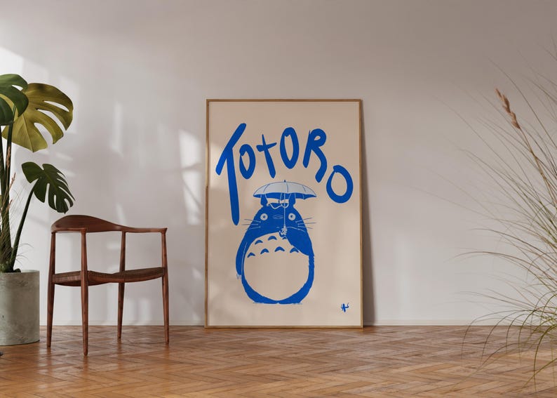 K&ouml;nnte beinhalten: Ein gerahmtes Poster mit einer blauen Illustration von Totoro mit einem Regenschirm und dem Wort "Totoro" in blauer Schrift. Das Poster steht vor einem neutralen Hintergrund, mit einem Holzstuhl und einer Topfpflanze.