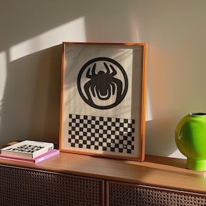 May include: A framed art print featuring a black spider emblem within a circle, above a black and white checkered pattern. The artwork is displayed on a wooden cabinet, alongside books and a green vase.