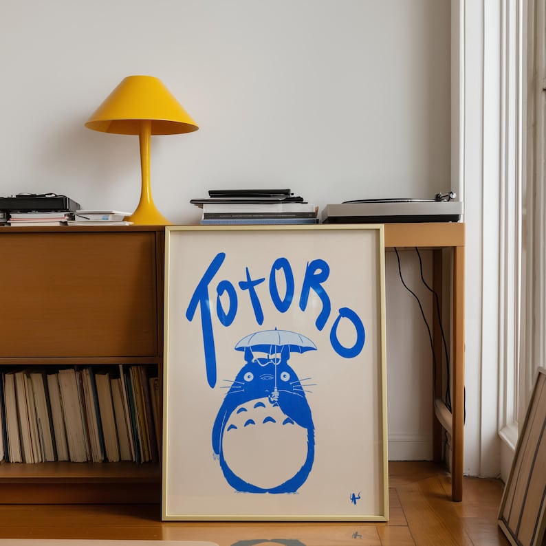 K&ouml;nnte beinhalten: Ein gerahmter Kunstdruck mit der Figur Totoro in Blau, die einen Regenschirm h&auml;lt, mit dem Wort "Totoro" dar&uuml;ber. Der Druck hat einen cremefarbenen Hintergrund und einen goldenen Rahmen. Eine gelbe Lampe und ein Holzschrank sind im Hintergrund.