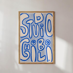 May include: A framed art print with the words "STUDIO GILI" in blue, stylized lettering on a light blue background. The artwork is in a wooden frame and hangs on a white wall.