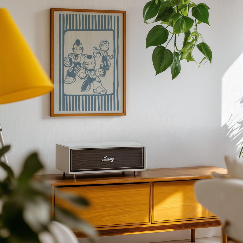 May include: A white and brown speaker, branded "Jimmy", rests on a wooden cabinet. A framed print featuring robot illustrations hangs on the wall. A yellow lamp and a hanging plant are also visible.