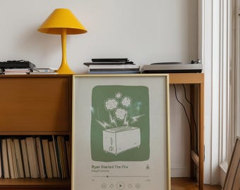 May include: A framed art print with a green background and a white illustration of a toaster with lightning bolts and clouds. The text "Ryan Started The Fire" is below the image. A yellow lamp and a wooden cabinet are in the background.