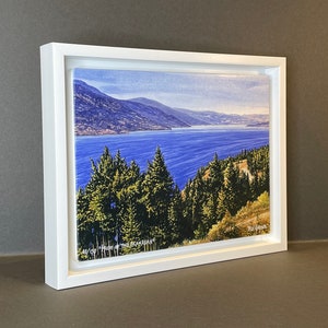 Peut inclure: Une impression encadrée d'un paysage intitulé "Pride of the Okanagan", représentant un lac entouré de montagnes et de conifères. L'impression est dans un cadre blanc et est numérotée 48/150.