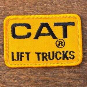 May include: A rectangular yellow patch with black embroidered text. The text reads "CAT" above "LIFT TRUCKS". The "CAT" text has a registered trademark symbol. The patch has a yellow stitched border.