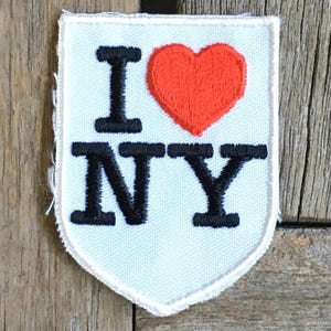 Puede incluir: Parche bordado con la frase "I ❤️ NY" en letras negras y un corazón rojo sobre un fondo blanco en forma de escudo. El parche tiene un borde blanco y está sobre una superficie de madera.