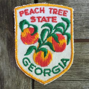 May include: Embroidered patch featuring the words "PEACH TREE STATE GEORGIA" in red and green. The patch has a white background with three peaches in the center, each with red and yellow coloring and green leaves. The patch is bordered with a yellow outline.