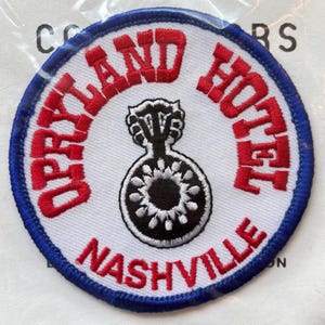 May include: A circular patch with a white background, a blue border, and red lettering. The text reads "OPRYLAND HOTEL NASHVILLE". A black and white guitar-shaped design is in the center.