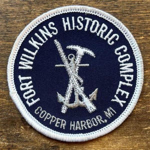 Może przedstawiać: Okrągła naszywka w kolorze granatowym z białym haftowanym tekstem i szczegółowym srebrnym emblematem. Tekst brzmi "FORT WILKINS HISTORIC COMPLEX COPPER HARBOR, MI". Emblemat przedstawia skrzyżowane narzędzia i kotwicę.