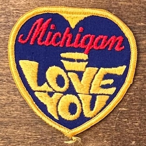Puede incluir: Parche en forma de corazón con las palabras "Michigan LOVE YOU" bordadas en rojo y amarillo sobre fondo azul. El parche tiene un borde amarillo, ideal para una chaqueta o bolso.