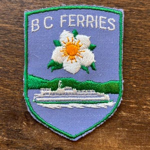 May include: Embroidered patch with a shield shape, featuring the text "BC FERRIES" in white. The patch has a light blue background with a white flower and a ferry boat design. The border is green.