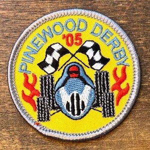 May include: A circular embroidered patch with a yellow background featuring a blue race car, two black and white checkered flags, and red flames. The text reads PINEWOOD DERBY and '05 in blue and red thread, finished with a silver-grey stitched border.