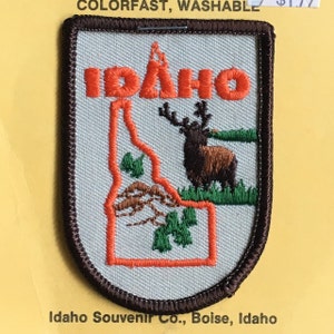 Può includere: Una toppa ricamata bianca con un bordo marrone. La toppa presenta un contorno dello stato dell'Idaho con la parola "IDAHO" in lettere arancioni sopra lo stato. All'interno del contorno dello stato c'è un alce marrone con corna e fogliame verde. Il testo "Idaho Souvenir Co., Boise, Idaho" è stampato sotto la toppa.