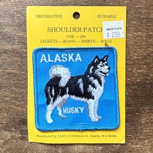 Pode incluir: Remendo de ombro quadrado azul com um cão Husky preto e branco, as palavras "ALASKA" e "HUSKY". O remendo está em um suporte amarelo com as palavras "DECORATIVE" e "DURABLE".