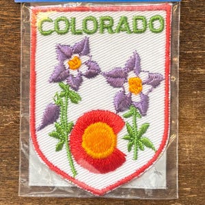 May include: Embroidered patch featuring the word "COLORADO" in green above a design of purple and yellow flowers, a red and orange "C" shape, and green stems and leaves. The patch has a white background and a red border.