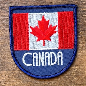 May include: Embroidered patch featuring the Canadian flag. The design includes a red maple leaf on a white background, flanked by red stripes, with the word "CANADA" in white lettering on a blue border.