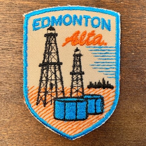 May include: A beige patch with a blue border featuring the word "EDMONTON" above "Alta" in orange script. The patch depicts two black oil derricks, blue tanks, and an orange and blue skyline.