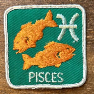 May include: A square embroidered patch with a green background and a white border. It features two orange fish swimming in opposite directions, a light blue Pisces zodiac symbol, and the word PISCES embroidered in white capital letters below.
