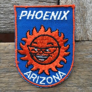 May include: Embroidered patch featuring a smiling sun wearing glasses, with the text "PHOENIX ARIZONA" in white lettering on a blue background. The sun is orange with black outlines and details. The patch has a blue border.