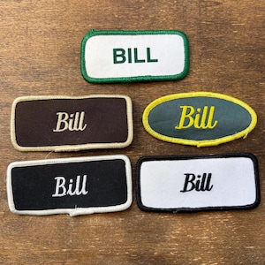 May include: Five embroidered name patches with the name "Bill". The patches are in various colors, including white, black, brown, and green, with contrasting border stitching. The text is in a simple, bold font.