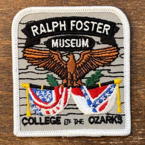 May include: Square patch with a gray background and white border. The patch features the text "RALPH FOSTER MUSEUM" above a brown eagle with outstretched wings. Below the eagle are two flags and the text "COLLEGE OF THE OZARKS."