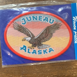 May include: A red, white, and blue woven patch with a bald eagle flying over a sunset. The patch says "Juneau, Alaska" in a teal blue font.
