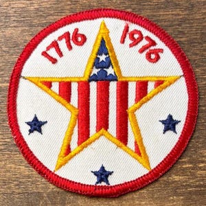 Peut inclure: Écusson circulaire avec une bordure rouge et les années "1776" et "1976" en rouge. Il présente une grande étoile jaune avec des rayures rouges et blanches, une étoile bleue au centre et trois étoiles bleues autour.