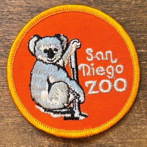 May include: Embroidered patch featuring a koala bear clinging to a branch, set against an orange background. The text "San Diego ZOO" is written in white script. The patch has a yellow stitched border.
