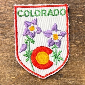 May include: A shield-shaped embroidered patch with a white background and a red border. The word COLORADO is stitched in green at the top. The design features two purple columbine flowers with yellow centers and a red and yellow circular emblem.